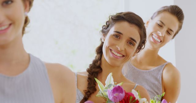 Three young bridesmaids are standing in a line, smiling and holding colorful bouquets in a bright, sunlit room. Their positive expressions and elegant dresses suggest they are part of a joyous wedding celebration. This image is ideal for use in wedding invitations, bridal magazines, event planning promotions, and blog posts about weddings and bridal parties.