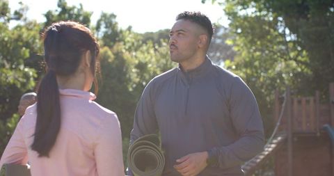 Male fitness coach holding mat coaching woman outdoors at park playground under sunlight