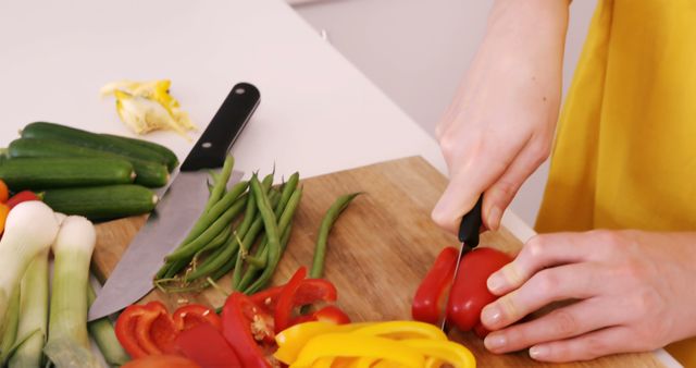 Hands slicing red and yellow bell peppers on a wooden cutting board alongside various other fresh vegetables such as leeks, green beans, zucchini, and knife. Ideal for content related to healthy eating, food preparation, cooking tutorials, recipes, and kitchen activities.