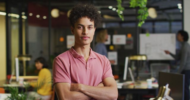 Confident young man with arms crossed in a modern office setting, with colleagues collaborating in the background. Perfect for business, teamwork, and leadership concepts.