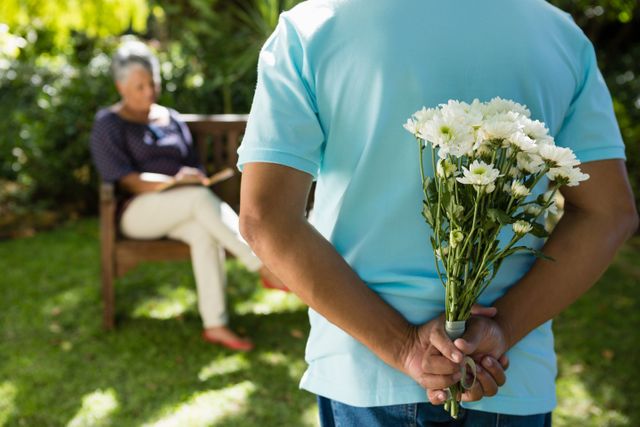 Senior man holding a bouquet of flowers behind his back, preparing to surprise a woman sitting on a bench in a garden. Ideal for themes of romance, elderly love, surprises, and outdoor activities. Can be used in advertisements, greeting cards, or articles about relationships and senior lifestyles.