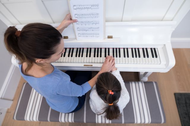 Mother guiding daughter in playing piano at home, emphasizing family bonding and music education. Ideal for use in articles about parenting, music lessons, home education, and family activities.