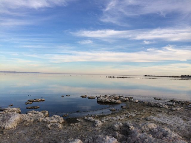 This image shows a calm and tranquil coastal scene with serene reflections of the cloudy sky on the peaceful water. The foreground features a rocky shore, adding a rugged texture to the idyllic landscape. The expansive horizon enhances the sense of peace and natural beauty. Perfect for use in travel blogs, environmental campaigns, relaxation themes, or as a soothing background.