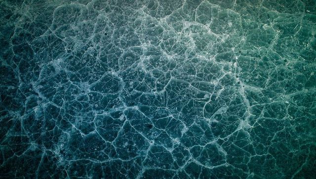 Top-down cracked ice expanse forming honeycomb fissures over frozen lake teal mosaic