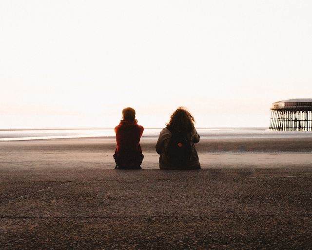 Pairs of friends sitting on a sandy beach near the pier during sunset, creating a serene and peaceful atmosphere. Perfect for themes of friendship, togetherness, relaxation, and holiday vibes. Can be used in travel blogs, friendship-themed advertisements, leisure activity promotions, and coastal environment features.