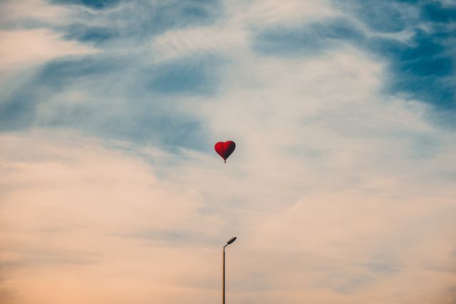 Red heart-shaped hot air balloon floating above white clouds against pastel blue and soft orange evening sky. Symbolizing love and romance, perfect for Valentine's Day themes, romantic ambiance, and adventure-related concepts.