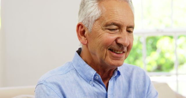 An elderly man smiling and dressed in a light blue shirt, indoors with natural light. Suitable for representations of aging, contentment, happiness, and relaxed lifestyle. Perfect for use in health and wellness articles, senior living advertisements, and family-oriented promotions.