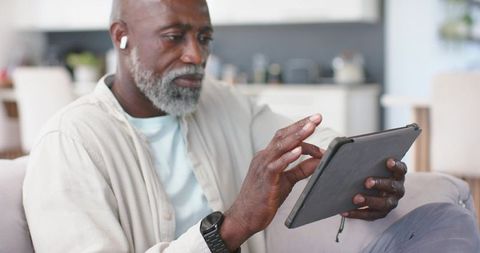 Senior African American man using tablet on sofa with earbuds in open-plan living room