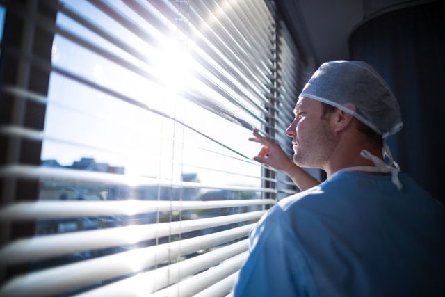 Doctor in scrubs looking through window with sunlight streaming in. Ideal for use in healthcare, medical, and hospital-related content, emphasizing contemplation, reflection, and the daily life of medical professionals.