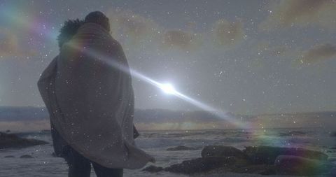 Romantic couple wrapped in blanket under starry sky at sea shore