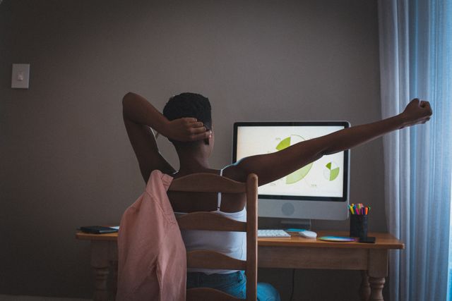African American woman stretching while working from home, looking at computer screen with charts. Ideal for illustrating remote work, home office setups, productivity tips, and work-life balance during quarantine or lockdown periods.