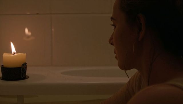 Woman sitting at bathtub edge gazing at candlelight reflection, moody evening solitude