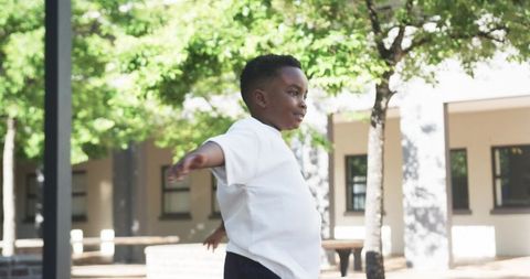 Joyful boy balancing with outstretched arms on sunlit courtyard playground among green trees