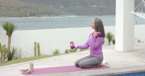 Senior woman kneeling lifting dumbbells on waterfront patio yoga mat overlooking sea