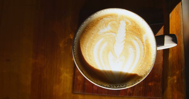 A cup of coffee with intricate latte art sits on a wooden surface, bathed in sunlight, with copy space. The warm lighting accentuates the creamy texture of the foam, inviting a moment of relaxation with a hot beverage.