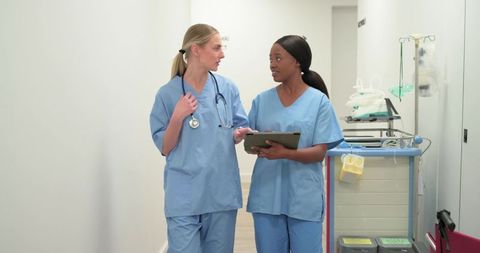 Diverse female nurses walking, discussing patient care and holding tablet in hospital corridor
