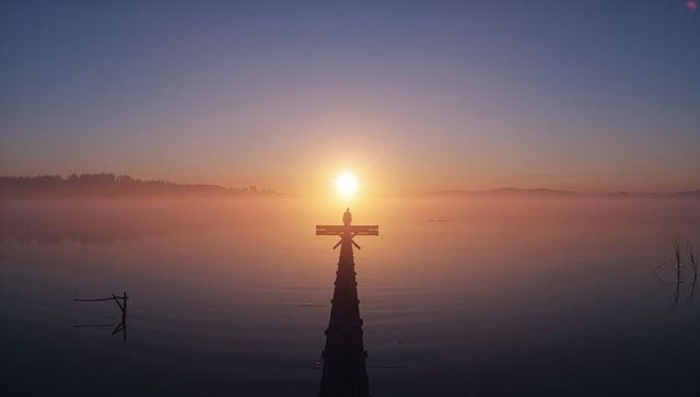 Silhouetted figure standing on jetty watching misty sunrise over lake with cross platform