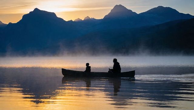 Paddling canoe at dawn on misty mountain lake, parent and child silhouette