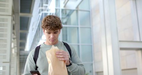 Young commuter checking smartphone while holding sandwich with backpack in modern glass atrium