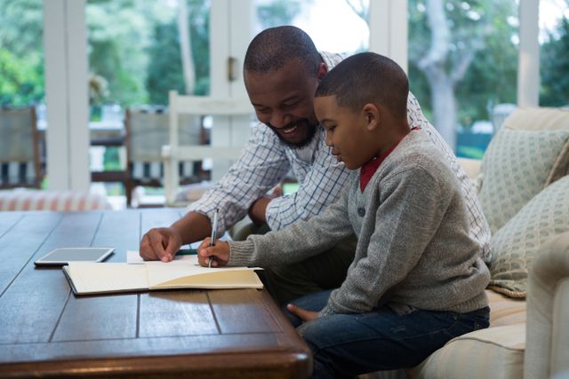 Father and son sitting together in a cozy living room, father helping son with homework. Ideal for use in educational materials, parenting blogs, family-oriented advertisements, and articles about fatherhood and childhood learning.