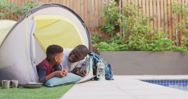 Father spending quality time with son in a backyard camping environment, inside opened tent. Use for themes related to family bonding, father-child relationships, outdoor activities, summer fun, parenting, and recreational activities.