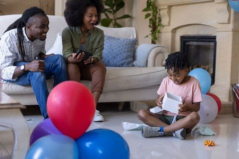 African american parents celebrating child opening gift at home with balloons