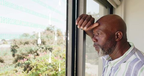 Contemplative senior man gazing through window with translucent binary data overlay at home