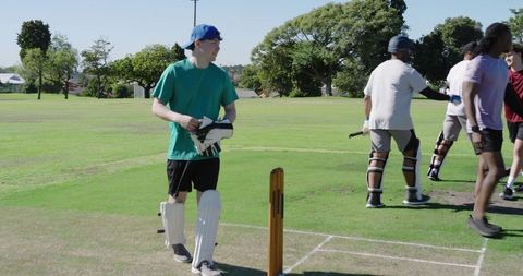 Young male batsman walking off crease wearing pads and gloves near wooden stumps in park