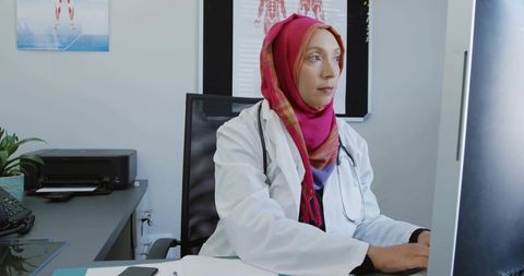 Asian female doctor wearing hijab typing on computer in clinic with stethoscope, charts
