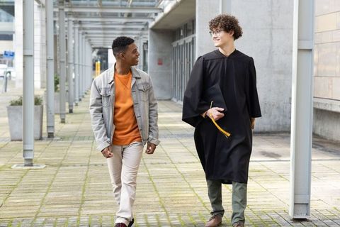 Graduating friends walking and talking on campus walkway wearing cap and gown
