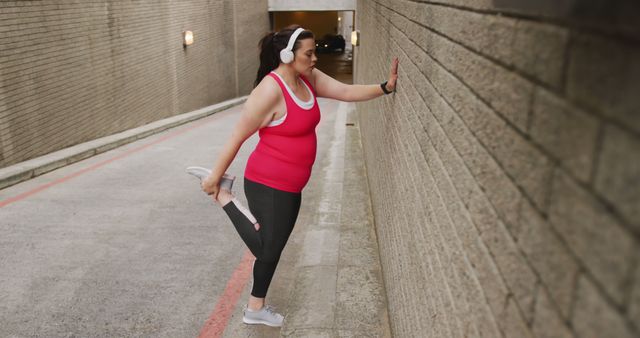 Woman wearing red top and gray leggings stretching her leg against a brick wall outdoors, listening to music with headphones. Suitable for topics like health, fitness, outdoor exercise routines, and active lifestyle promotions.