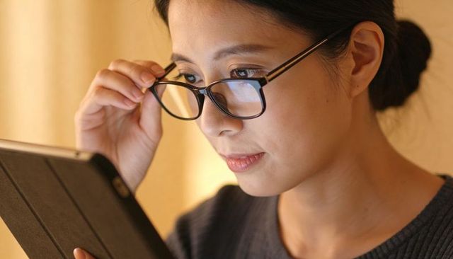 Asian woman reading tablet while adjusting glasses with screen reflection closeup warm light