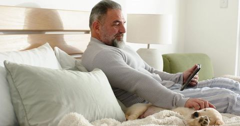 Senior man reclining in bed holding tablet and petting sleeping puppy in morning light