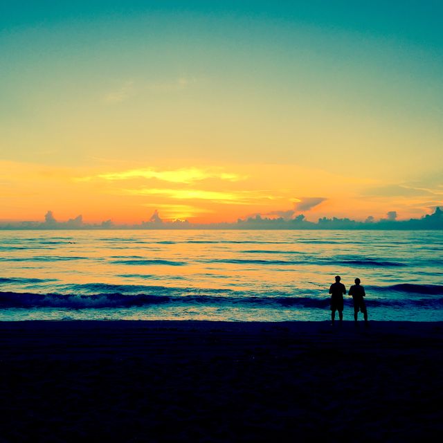Two people standing at ocean shore watching a vibrant sunset. Ideal for themes of love, tranquility, travel, nature, romance, and togetherness. Can be used in advertisements, travel brochures, romantic cards, inspirational posters, websites on coastal vacations, and peaceful lifestyle blogs.
