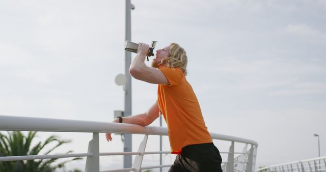 This photo shows a young blonde man wearing an orange shirt, leaning on a railing and drinking from a water bottle after exercising. The clear sky and sunlight create a bright and energetic atmosphere. Perfect for uses in fitness-related promotions, exercise guides, outdoor activity advertisements, and health and wellness articles emphasizing the importance of hydration and outdoor workouts.