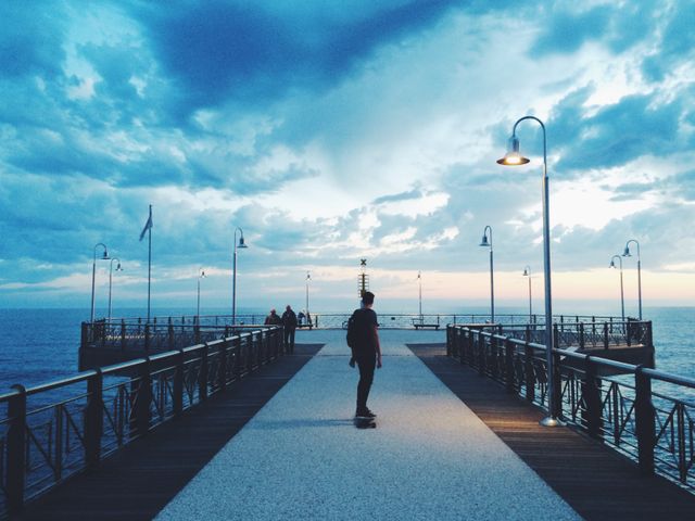 Teenager skateboarding on a pier during sunset with a cloudy sky. Ideal for themes of freedom, teenage adventure, or tranquility. Suitable for travel blogs, lifestyle promotions, or inspirational content.