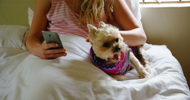 Caucasian female teenager petting her little dog and using smartphone on bed at home. Domestic life, technology, pets, animals and care.