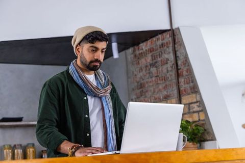 Indian man working on laptop in loft kitchen with exposed brick wall and coffee mug
