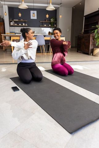 Two women practicing yoga and stretching shoulders on mats in bright home living room