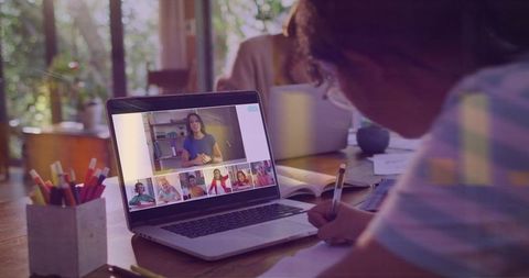 Child studying and writing during online classroom with teacher on laptop screen