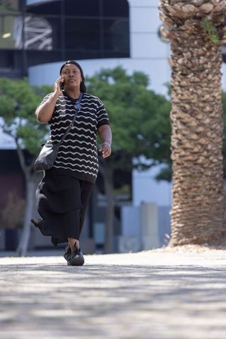 Urban woman walking and talking on phone in sunlit plaza with crossbody bag