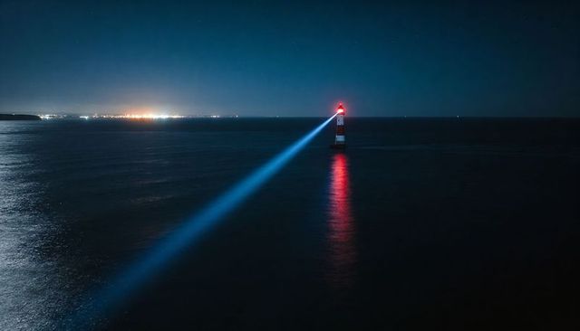 Lighthouse beacon casting blue beam across dark sea with red glow and harbor lights