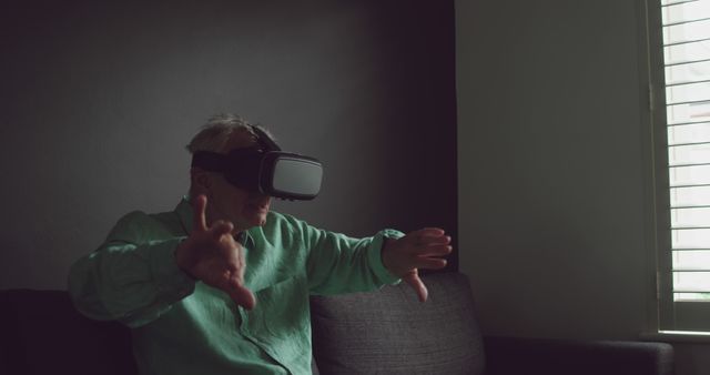 Senior man engaging with virtual reality using a VR headset, gesturing with hands in a dimly-lit room with partial window lighting. Ideal for illustrating technology adoption among older adults, promoting virtual reality experiences for seniors, and showcasing the use of VR for innovative applications.