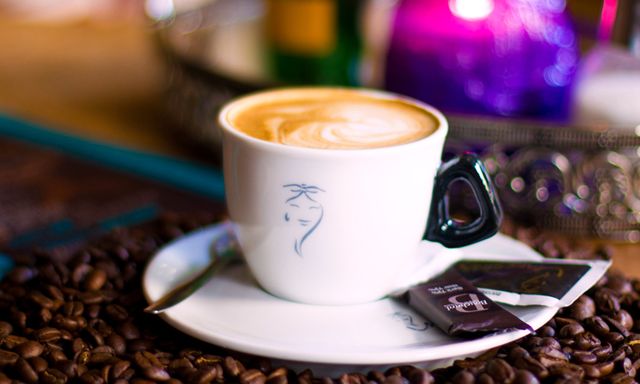 Cappuccino with latte art served in white cup and saucer on table covered with coffee beans. Packet and spoon are on the saucer. Ideal for coffee shop advertisements,-food blogs, cafe menu displays, barista training materials, or social media promotions for coffee lovers.