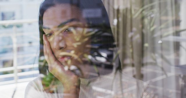 Young woman with a hand on her face, looking out the window with a thoughtful expression. The reflection of plants and surroundings creates a serene and contemplative mood. Useful for mental health contexts, articles on introspection, or advertisements aiming to convey thoughtfulness.
