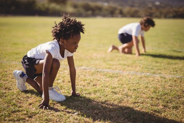 Schoolgirls are preparing for a race in a park, crouched in the ready position on a sunny day. This image can be used for educational materials, sports and fitness promotions, youth activities, and teamwork concepts.
