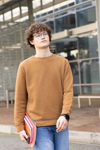 Teen student walking on campus wearing camel pullover and glasses holding colorful notebooks