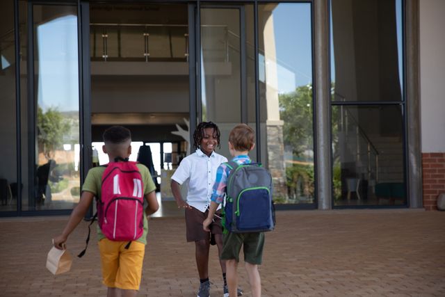 Three multiracial elementary schoolboys walking towards school entrance, smiling and carrying backpacks. Ideal for use in educational materials, back-to-school promotions, advertisements for school supplies, and articles about childhood friendships and diversity in education.