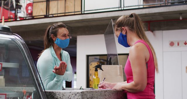 Two women wearing sporty attire and protective masks have an interaction at a gym reception desk. One woman is behind the desk checking in or signing something, while the other casually converses with her. The setting appears to be modern and well-equipped gym or fitness center. Useful for topics related to fitness during the pandemic, safety measures in public places, health and wellness industries.