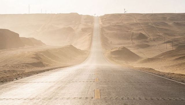 Endless desert highway climbing toward hazy horizon with gentle dunes and utility poles